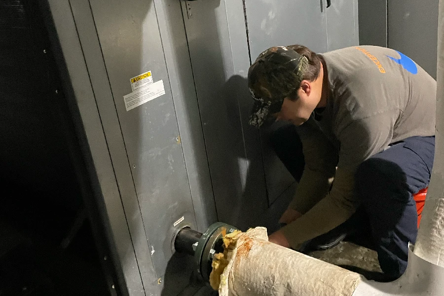 A person kneels beside industrial machinery, performing maintenance on a metal shaft with tools and a protective cloth covering part of the equipment.