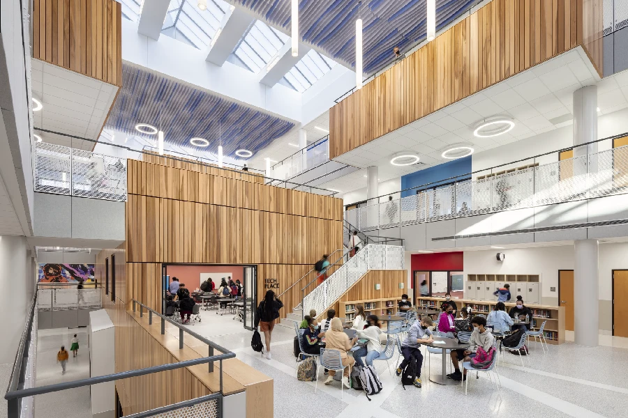 A spacious, modern school atrium with wood paneling, large skylights, students seated at tables, and others walking or talking in a bright, open environment.