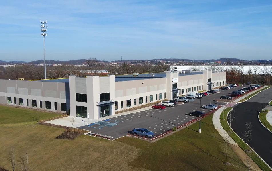 A large, two-story commercial building with a parking lot partially filled with cars, surrounded by grass and trees under a clear sky.