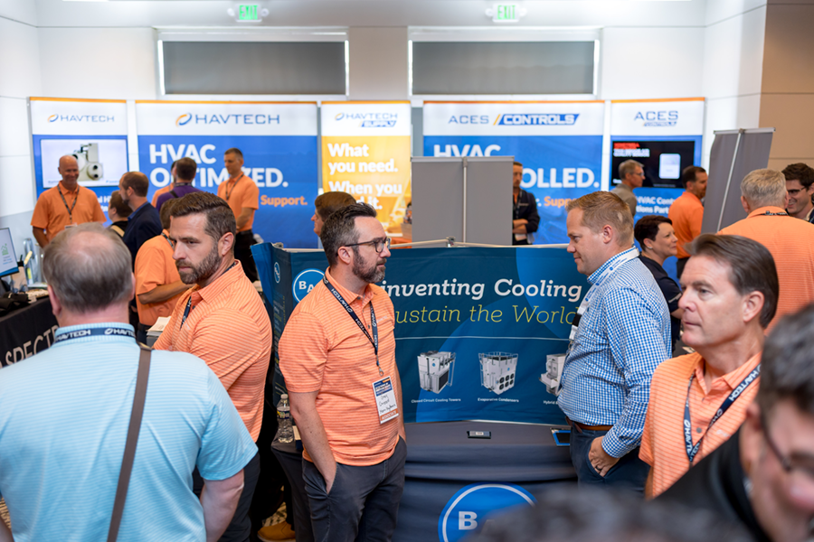 Three men stand at a trade show booth discussing HVAC equipment; brochures and machinery are displayed on the table in front of them.