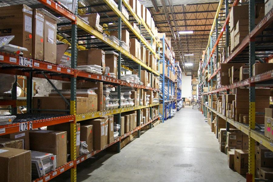 A warehouse aisle lined with tall metal shelves filled with cardboard boxes and various packages, with industrial lighting overhead.