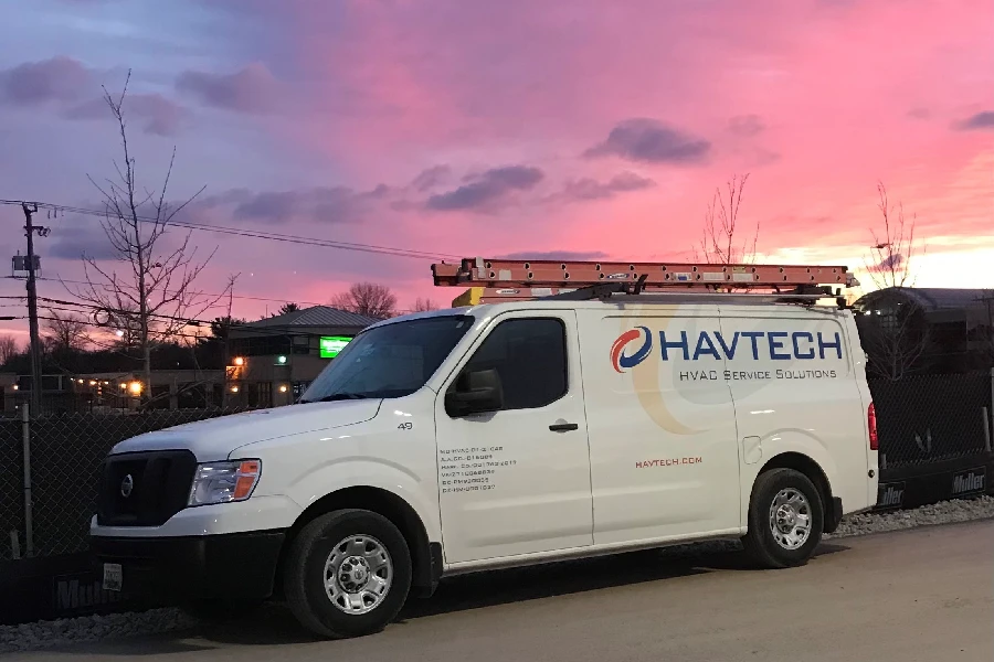 A white HAVTECH HVAC service van with a ladder on top is parked by a fence at sunset, with a pink and purple sky in the background.