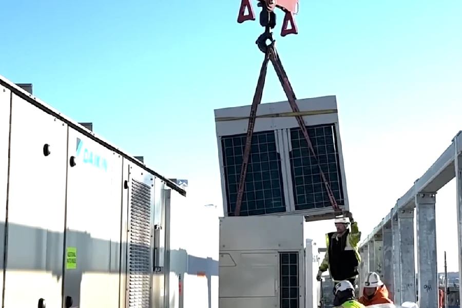 A construction crane lifts a large HVAC unit while workers in safety gear guide its placement between buildings on a sunny day.