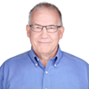 Older man with short gray hair and glasses smiles at the camera, wearing a blue button-down shirt, shown against a plain white background.