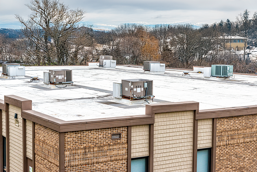 A snowy commercial building roof with several HVAC units and vents, surrounded by trees and a cloudy sky in the background.