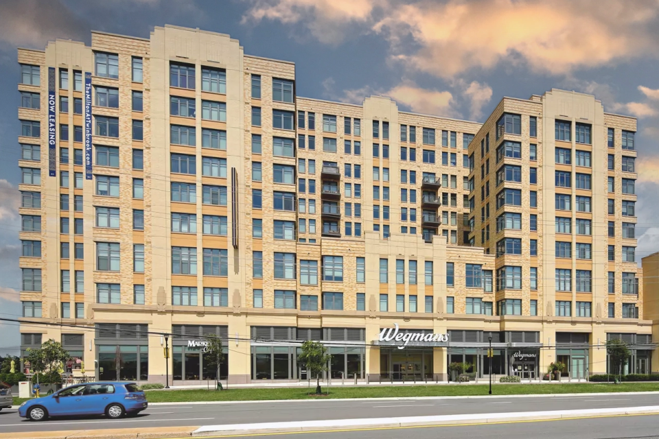 A multi-story tan brick apartment building with retail stores, including Wegmans, on the ground floor. A blue car is parked on the street in front. The sky is partly cloudy.