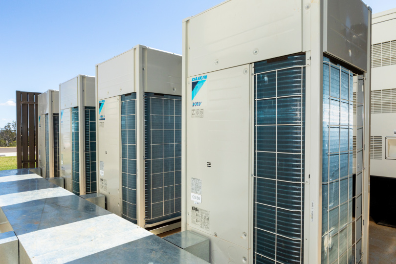 Several large Daikin outdoor air conditioning units, part of A2L VRF systems in hotels and multifamily buildings, are lined up next to a building on a sunny day.