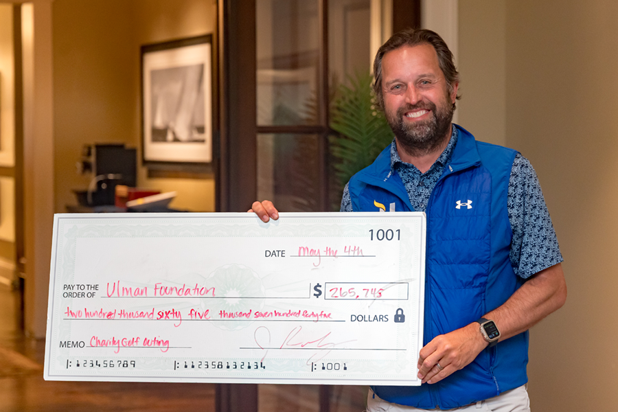 A man stands indoors holding a large ceremonial check for $265,745 to the Ulman Foundation, dated May 4th, raised through the 2026 Havtech Charity Golf Outing.