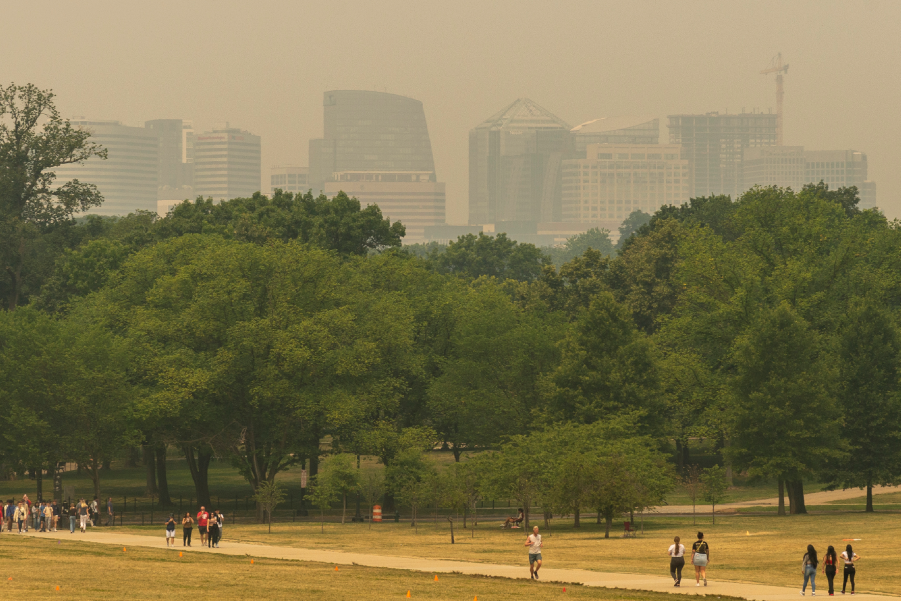 People walk along a path in a park with trees, while a city skyline is partially obscured by wildfire smoke in the background, highlighting growing outdoor and indoor air quality concerns.