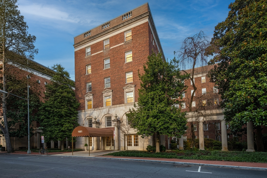 A multi-story red brick building with large windows, a tan entrance canopy, and trees surrounding the front, 1661 Crescent Place Washington DC is viewed from across the street.